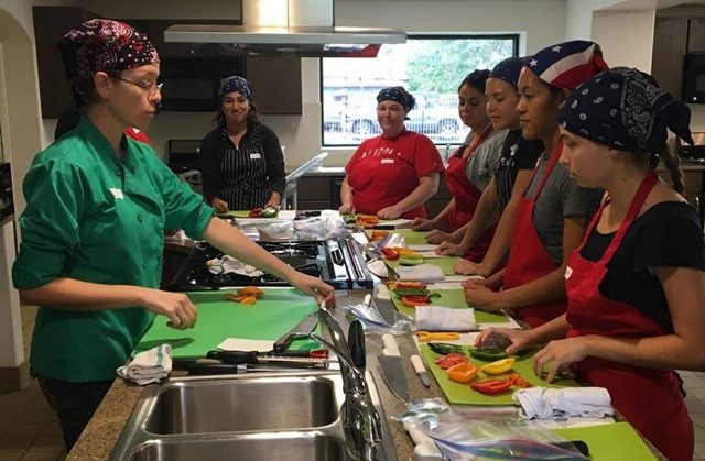 A cooking class stands around a countertop in a demonstration kitchen. The instructor stands on the left while 6 students stand attentively to the right, each with a cutting board and a variety of vegetables in front of them.